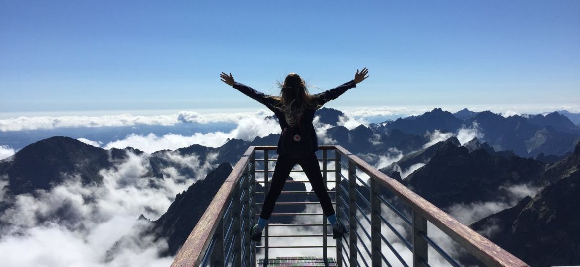 person standing on hand rails with arms wide open facing the mountains and clouds